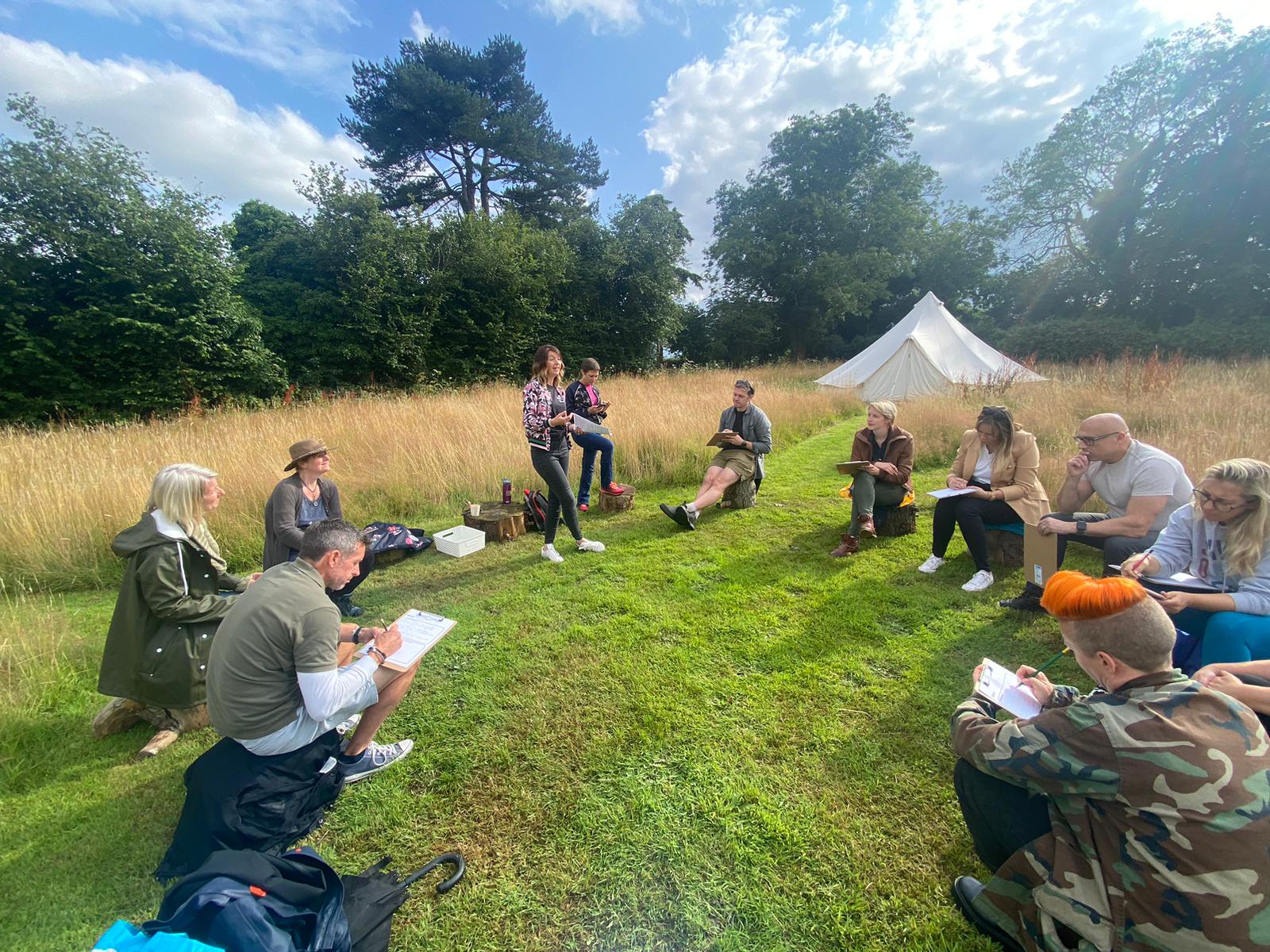 A group sitting on logs in long grass for an outdoor workshop, with a bell tent in the background.